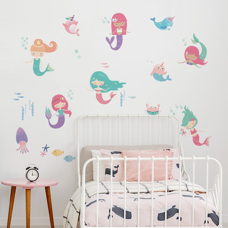 Removable Walls Sticker Mermaids with two narwhals, a crab, a squid and some fish, in bright vibrant colours, as seen in the small option, above a white metal frame bed, with a pink and navy whale-themed bedspread and pillow case set. Next to the bed, is a pink round bedside table, with three light brown legs, and an alarm clock on top.