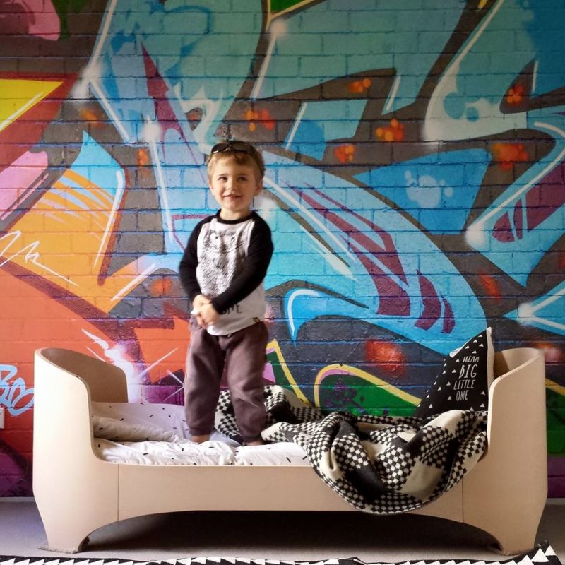 Little boy stands on his bed in front of a wall with graffiti on it