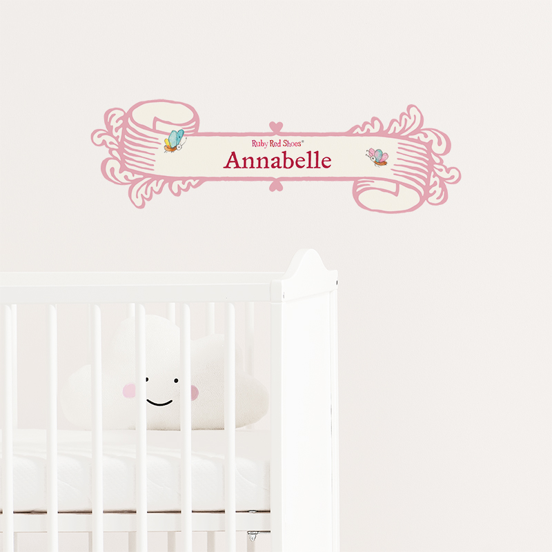 Customisable Ruby Red Shoes Name Banner in pink with the name 'Annabelle' on an example wall behind a white wooden cot with a white cloud-shaped solid cushion, a cute smiling face printed on it looking out at the viewer. Ruby Red Shoes By Kate Knapp.